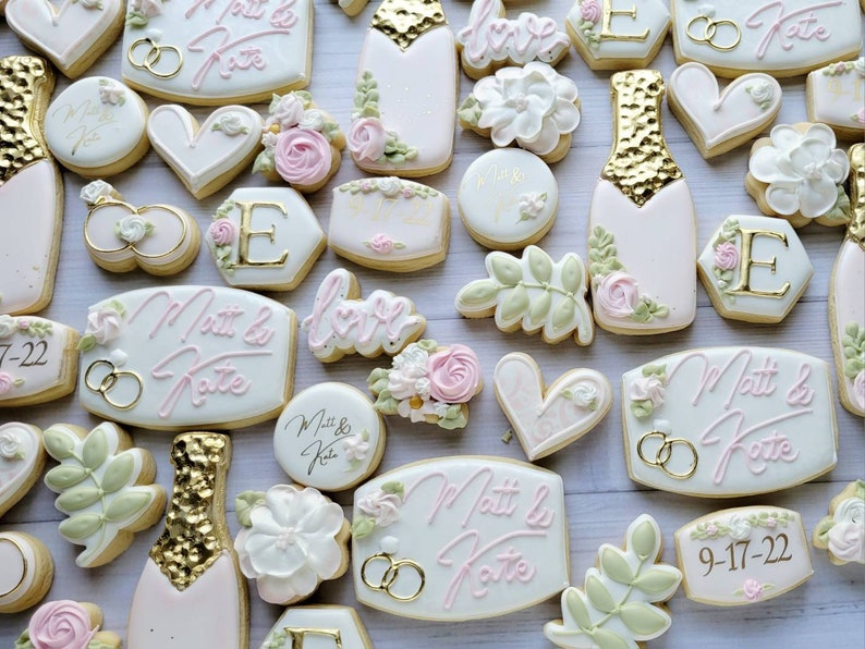Pu&ograve; includere: Una collezione di biscotti decorati con bottiglie di champagne con foglia d'oro, fiori bianchi, foglie verdi, cuori e targhe esagonali. Diversi biscotti mostrano il testo Matt & Kate e la data 9-17-22 in glassa rosa e dorata.