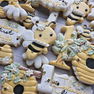 May include: A collection of decorated sugar cookies featuring yellow and black bees, yellow beehives with green leaves and white flowers, white daisies, and the number one. Some cookies display the text "Juliana's first bee-day" in black and gold.
