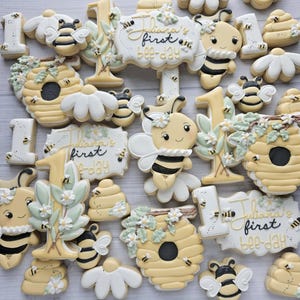 May include: An assortment of decorated sugar cookies with a bee theme. The cookies include bees, beehives, daisies, the number 1, and signs that read "Juliana's first bee-day". The cookies are primarily yellow, white, and black.