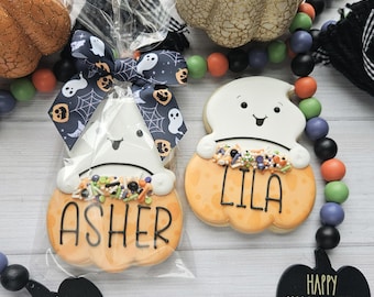 May include: Two ghost-shaped sugar cookies decorated with white icing, black facial features, and orange pumpkin bases. One cookie is in a clear plastic bag with a ghost and spiderweb ribbon. The cookies feature the names ASHER and LILA.