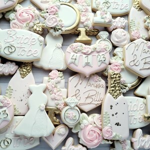 Pu&ograve; includere: Una collezione di biscotti decorati a tema matrimonio, incluse forme di abiti, bottiglie di champagne, cuori e anelli. I biscotti sono glassati in bianco, rosa pallido e oro, con il testo Bride to be e Marissa & Billy.