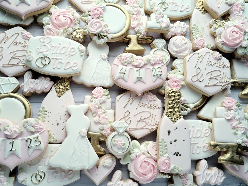 Pu&ograve; includere: Una collezione di biscotti decorati a tema matrimonio, incluse forme di abiti, bottiglie di champagne, cuori e anelli. I biscotti sono glassati in bianco, rosa pallido e oro, con il testo Bride to be e Marissa & Billy.