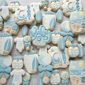 Puede incluir: Una colecci&oacute;n de galletas decoradas en tonos azul claro, blanco y beige. Las formas incluyen bodies de beb&eacute; con el texto 'crawl. walk. surf.', biberones, tablas de surf, sonajeros, cochecitos y escenas de playa con 'Baby on Board'.