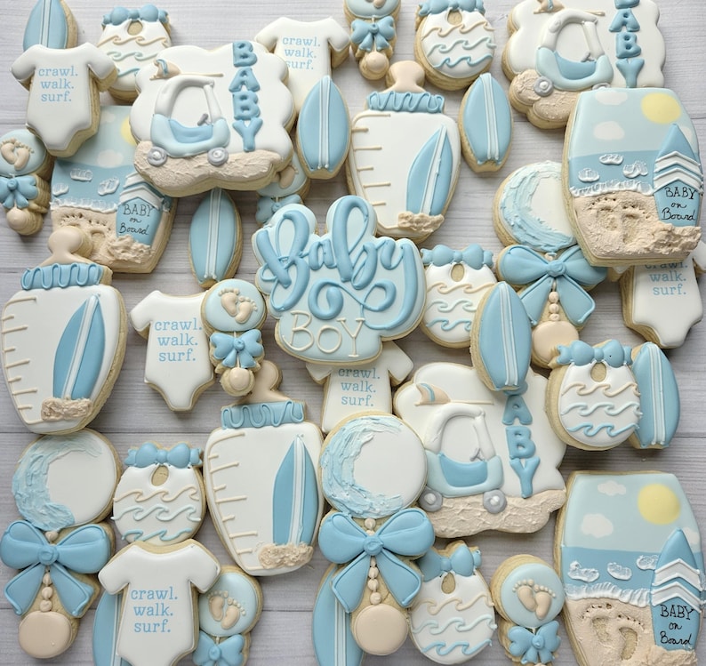 Puede incluir: Una colecci&oacute;n de galletas decoradas en tonos azul claro, blanco y beige. Las formas incluyen bodies de beb&eacute; con el texto 'crawl. walk. surf.', biberones, tablas de surf, sonajeros, cochecitos y escenas de playa con 'Baby on Board'.