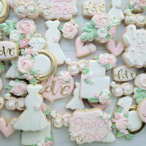 Pu&ograve; includere: Una collezione di biscotti a tema matrimonio decorati con glassa bianca, rosa e verde. Le forme includono abiti, cuori, anelli con diamante, fedi nuziali e torte, con alcune decorazioni che riportano la scritta dorata 'love' o 'bride to be'.