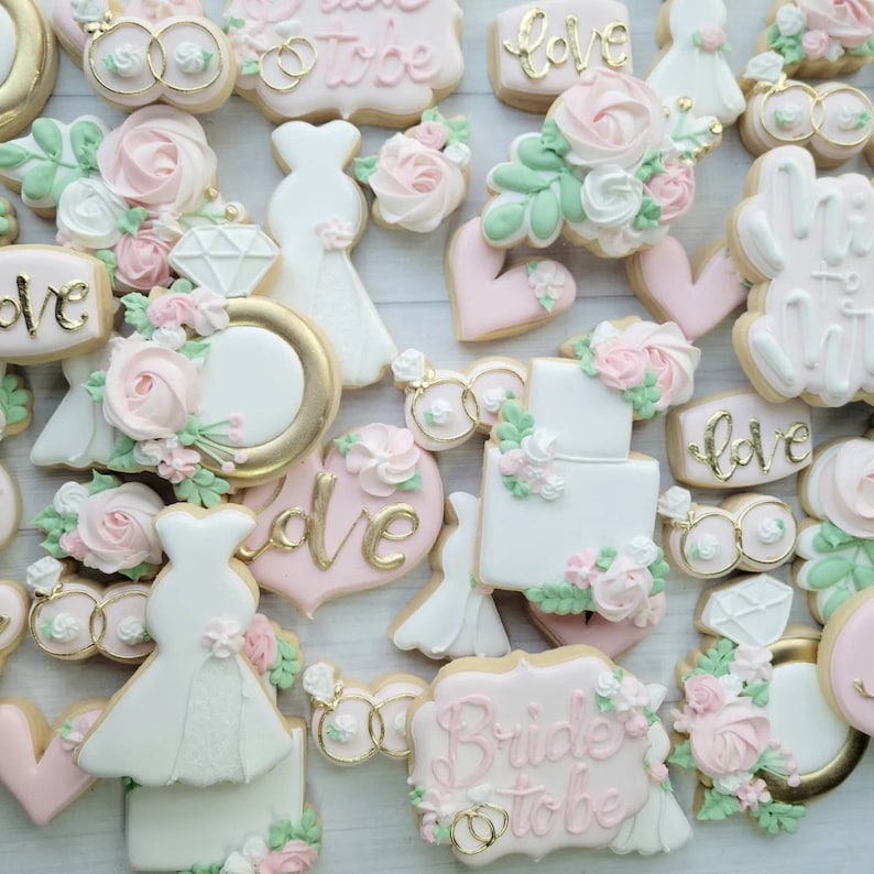 Pu&ograve; includere: Una collezione di biscotti a tema matrimonio decorati con glassa bianca, rosa e verde. Le forme includono abiti, cuori, anelli con diamante, fedi nuziali e torte, con alcune decorazioni che riportano la scritta dorata 'love' o 'bride to be'.