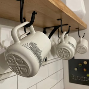 May include: Four white ceramic mugs with black handles hang from hooks on a wooden shelf. Each mug has a black and white graphic of a building on one side and a black and white logo on the other side.