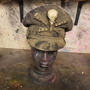 May include: A dirty, dark brown military style hat with a skull and crossbones design. The hat is made of leather and has a wide brim. The hat is sitting on a mannequin head.