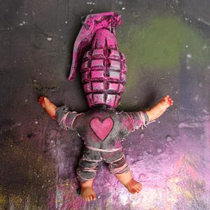 Könnte beinhalten: Ein einzigartiges Kunstwerk, das eine pinkfarbene Granate mit einem puppenartigen Körper kombiniert. Die Puppe hat ein graues Hemd mit einem rosa Herz und ausgestreckten Armen. Der Hintergrund ist eine dunkle Oberfläche mit bunten Farbspritzern.