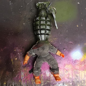 Könnte beinhalten: Ein einzigartiges Kunstwerk, das einen Granatenkopf mit einem Puppenkörper kombiniert. Die Granate ist dunkelgrau mit grünen Akzenten, während der Puppenkörper in grauen Stoff gekleidet ist. Die Gliedmaßen der Puppe sind hautfarben mit roten Akzenten.