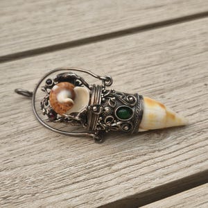 Puede incluir: Un colgante de plata con una intrincada jaula de filigrana metálica que sostiene una pequeña concha en espiral marrón y blanca. La pieza incluye una punta de concha blanca y bronceada, una piedra ovalada verde lisa y pequeños acentos rojos.