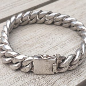 Peut inclure: Un bracelet gourmette de couleur argentée composé de maillons ovales épais et entrelacés, doté d'un fermoir rectangulaire à cliquet avec un petit loquet de sécurité intégré. La surface métallique présente une finition polie et usée.