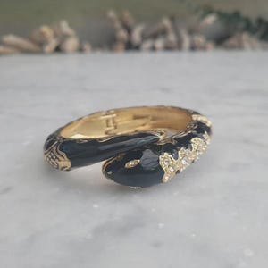 Peut inclure: Un bracelet jonc articulé doré en forme de serpent enroulé. Le bracelet présente une finition en émail noir brillant sur le corps et la tête, agrémenté de strass transparents étincelants sur le dessus et d'un mécanisme de charnière métallique.