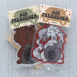 May include: Two air fresheners in clear packaging. One has a brown bear graphic, the other a white and gray cartoon animal with a turbocharger. The packaging reads "AIR FRESHENER" with "PINE" and "CHERRY" labels.