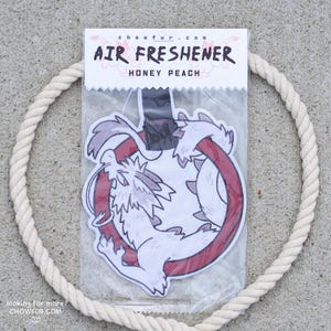 Puede incluir: Ambientador en un paquete de plástico transparente con un diseño de dragón de dibujos animados blanco y rojo. El texto "AIR FRESHENER HONEY PEACH" está impreso en la parte superior. Una cuerda blanca forma un círculo alrededor del ambientador.