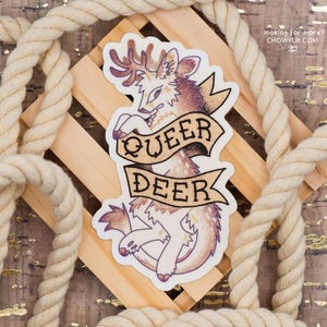 May include: A sticker featuring a deer illustration with the text "QUEER DEER" on a banner. The deer has brown and purple fur and antlers. The sticker is on a wooden background with a rope border. The text "looking for more? CHOWFUR.COM" is in the upper right corner.
