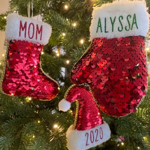 May include: Three red sequin Christmas ornaments with white fur trim. The ornaments are shaped like a stocking, a mitten, and a Santa hat. The stocking has the word "MOM" embroidered on it. The mitten has the word "ALYSSA" embroidered on it. The Santa hat has the year "2020" embroidered on it.