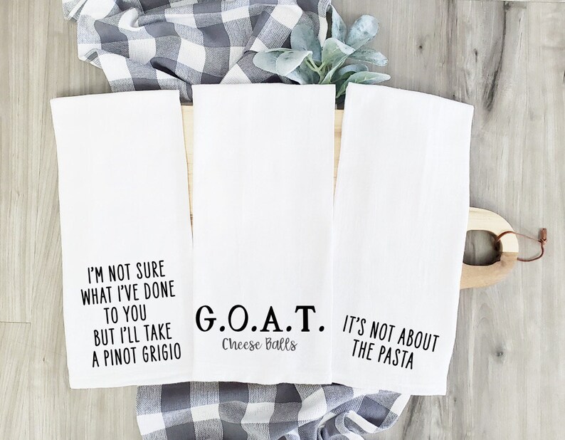 May include: Three white kitchen towels with black text. The first towel says "I'm not sure what I've done to you but I'll take a Pinot Grigio". The second towel says "G.O.A.T. Cheese Balls". The third towel says "It's not about the pasta".