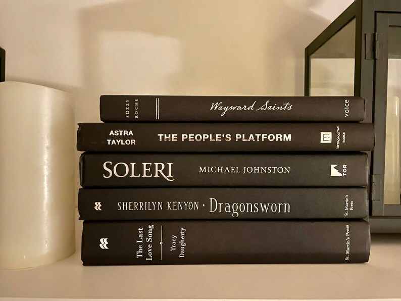 May include: A stack of five black hardback books with white text titles. The titles are "Wayward Saints", "The People's Platform", "Soleri", "Dragonsworn", and "The Last Love Song".