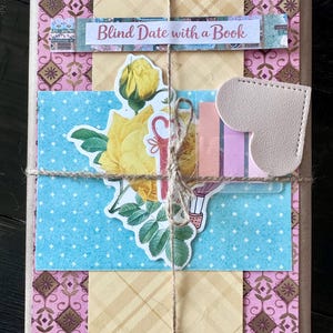 May include: A wrapped book with decorative paper and twine. The cover features a pink and brown floral pattern, with a blue polka dot section and yellow plaid paper. A sticker reads "Blind Date with a Book."