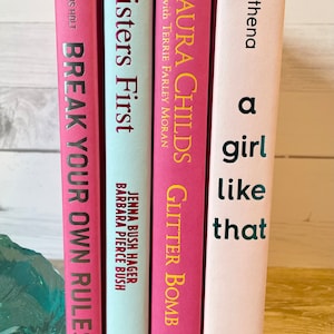 May include: A stack of five pink and light blue paperback books with titles including "Break Your Own Rules", "Sisters First", "Laura Childs Glitter Bomb", and "A Girl Like That".