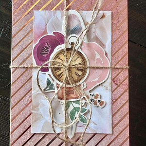 May include: A pink and gold striped gift box tied with twine. A decorative card with floral and clock stickers is attached. The card features pink and purple flowers and a vintage clock.