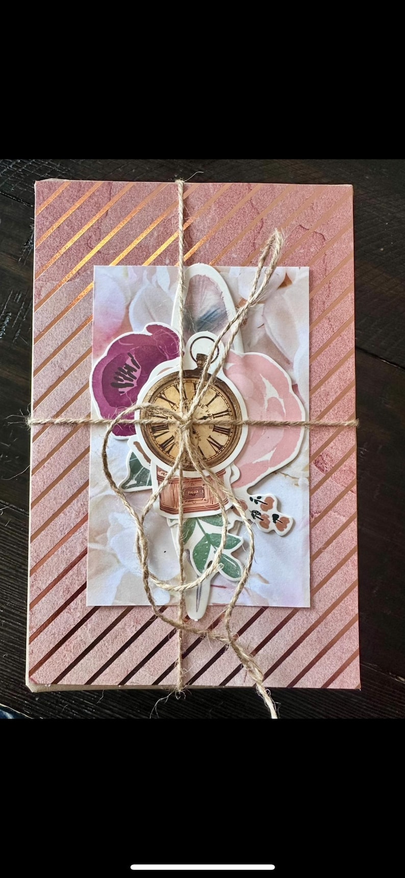 May include: A pink and gold striped gift box tied with twine. A decorative card with floral and clock stickers is attached. The card features pink and purple flowers and a vintage clock.
