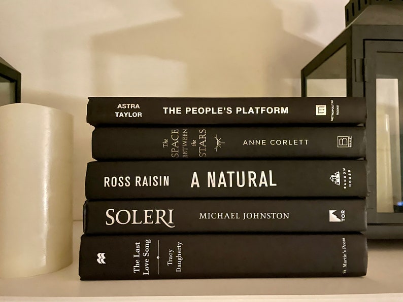 May include: A stack of five black hardback books with white text titles. The titles are "The People's Platform", "The Space Between the Stars", "A Natural", "Soleri", and "The Last Love Song".