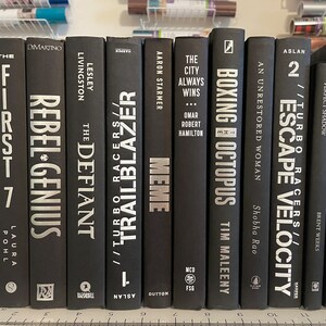 May include: A collection of ten black hardback books with silver lettering on the spines. The titles include "The First 7", "Rebel-Genius", "The Defiant", "Trailblazer", "Meme", "The City Always Wins", "Boxing Octopus", "Escape Velocity", "Turbo Racers", and "Ninja Future".
