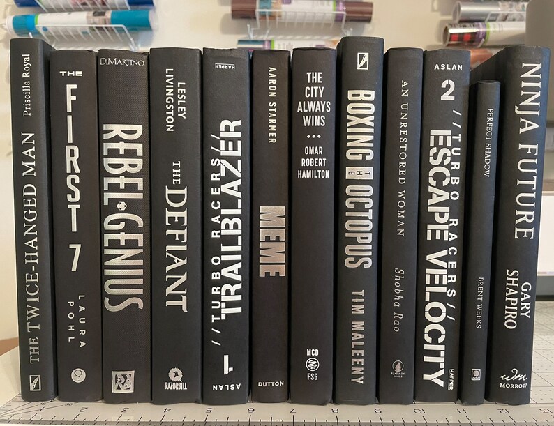 May include: A collection of ten black hardback books with silver lettering on the spines. The titles include "The First 7", "Rebel-Genius", "The Defiant", "Trailblazer", "Meme", "The City Always Wins", "Boxing Octopus", "Escape Velocity", "Turbo Racers", and "Ninja Future".