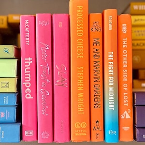 May include: A bookshelf with a variety of colorful books stacked on top of each other. The books have different titles and authors. Some of the books are yellow, orange, pink, and purple.