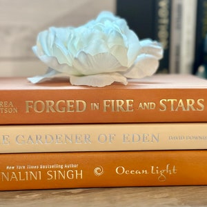 May include: A stack of three books with orange and cream covers. The top book is titled "Forged in Fire and Stars" by Andrea Robertson. The middle book is titled "The Gardener of Eden" by David Downie. The bottom book is titled "Ocean Light" by Nalini Singh.