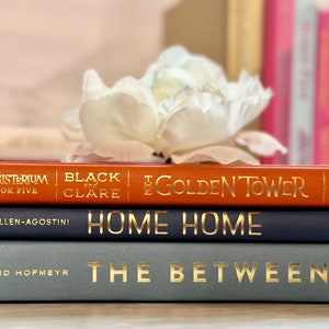 May include: A stack of five books with gold lettering on the spines. The top book is orange and says "The Golden Tower" and "Black by Clare". The second book is navy blue and says "Home Home" by "Lisa Allen-Agostini". The third book is light blue and says "The Between" by "David Hofmeyr".