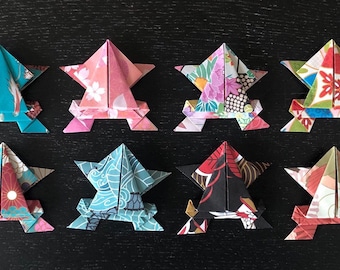 May include: Twelve origami frogs made from colorful patterned paper. The frogs are arranged in two rows of six, with each frog facing forward. The frogs are all the same size and shape, but each has a different pattern.