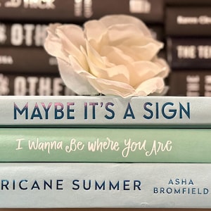 May include: A stack of three books with pastel covers. The top book reads "Maybe It's a Sign" in blue lettering. The middle book says "I Wanna Be where You Are" in cursive. The bottom book is titled "Hurricane Summer". A white flower rests on top.