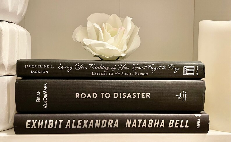 May include: A stack of three black hardback books with white titles. The top book is titled "Loving You, Thinking of You, Don't Forget to Pray: Letters to My Son in Prison" by Jacqueline L. Jackson. The middle book is titled "Road to Disaster" by Brian VanDemark. The bottom book is titled "Exhibit Alexandra Natasha Bell" by Crown.