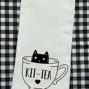 May include: White tea towel with a black and white illustration of a cat in a teacup. The text "Kit-Tea" is written below the cat.