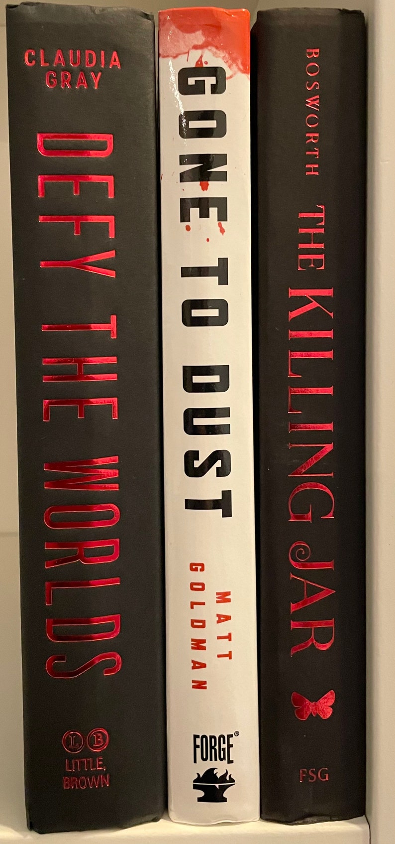May include: Three hardback books with black covers and red titles. The book on the left is "Defy the Worlds" by Claudia Gray, the middle book is "Gone to Dust" by Matt Goldman, and the book on the right is "The Killing Jar" by Bosworth.