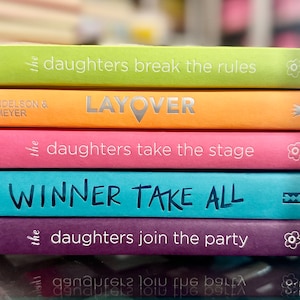 May include: A stack of five colorful paperback books with titles that include "the daughters break the rules", "LAYOVER", "the daughters take the stage", "WINNER TAKE ALL", and "the daughters join the party".