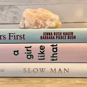May include: A stack of four books with different colored covers. The top book is light blue with the title "Sisters First" by Jenna Bush Hager and Barbara Pierce Bush. The second book is pink with the title "a girl like that" by bhathena. The third book is white with the title "Slow Man" by J.M. Coetzee. The bottom book is white with the title "Grand Central" by