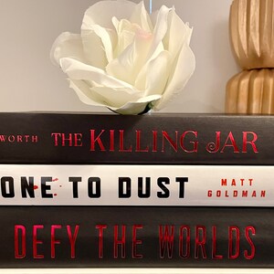 May include: A stack of three hardback books with black covers and red titles. The top book is titled "The Killing Jar" by Bosworth. The middle book is titled "Gone to Dust" by Matt Goldman. The bottom book is titled "Defy the Worlds" by Claudia Gray.