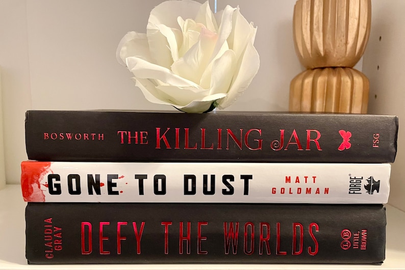 May include: A stack of three hardback books with black covers and red titles. The top book is titled "The Killing Jar" by Bosworth. The middle book is titled "Gone to Dust" by Matt Goldman. The bottom book is titled "Defy the Worlds" by Claudia Gray.