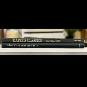 May include: Two black books stacked on a white shelf. The top book reads "KAFFE'S CLASSICS" in gold lettering, with "KAFFE FASSETT" and "Little, Brown" printed on the spine. The bottom book reads "Adam Elsheimer 1578-1610".