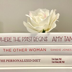 May include: A stack of three white books with red titles. The top book is titled "Where the Past Begins" by Amy Tan. The middle book is titled "The Other Woman" by Sandie Jones. The bottom book is titled "The Personalized Diet" by Eran Segal, PhD and Eran Elinav, MD, PhD. A white rose sits on top of the books.