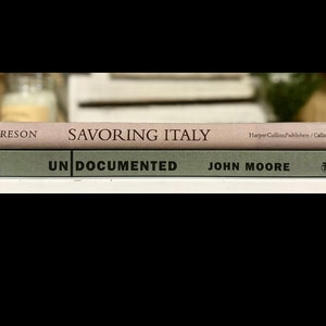 May include: Two books stacked on top of each other. The top book is pink and says "SAVORING ITALY" with the publisher "HarperCollins Publishers / Callaway" printed on the spine. The bottom book is green and says "UNDOCUMENTED" with the author "JOHN MOORE" printed on the spine.