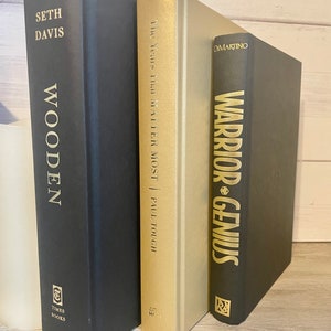 May include: Three hardcover books stacked on a wooden surface. The book on the left is black with gold lettering that reads "Seth Davis Wooden." The middle book is gold with black lettering that reads "The Years That Matter Most | Paul Touch." The book on the right is black with gold lettering that reads "DiMartino Warrior-Genius."