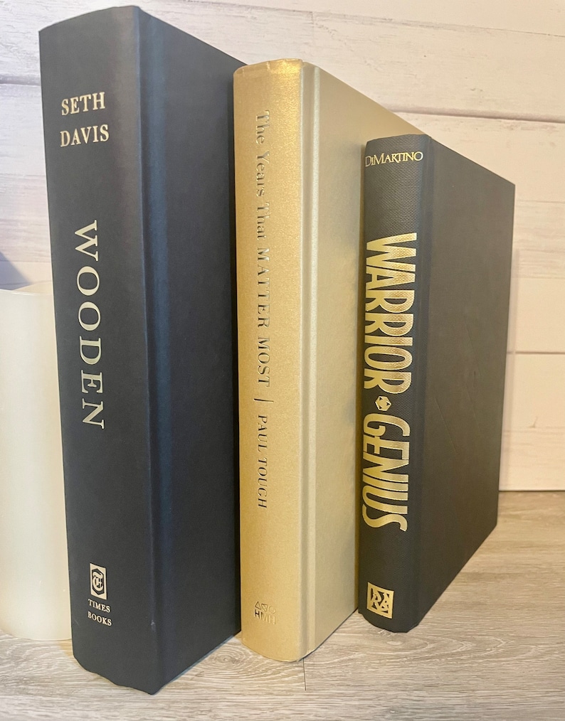 May include: Three hardcover books stacked on a wooden surface. The book on the left is black with gold lettering that reads "Seth Davis Wooden." The middle book is gold with black lettering that reads "The Years That Matter Most | Paul Touch." The book on the right is black with gold lettering that reads "DiMartino Warrior-Genius."