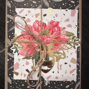 May include: A gift box wrapped in black paper with white feathers and arrow patterns. A floral design with pink peonies and gold accents is tied with twine. The box is on a dark surface.