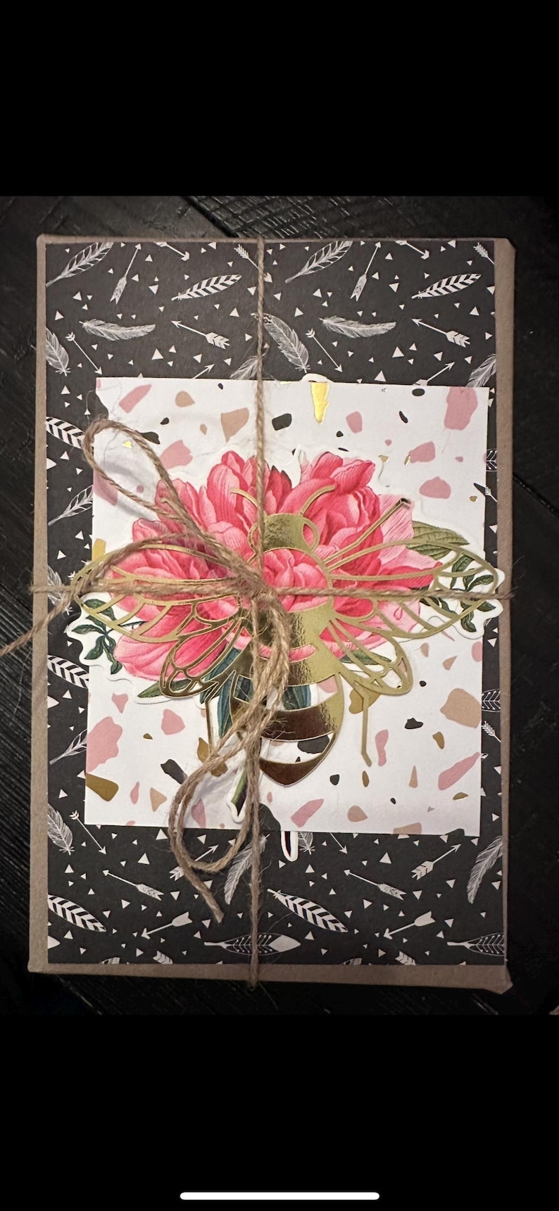 May include: A gift box wrapped in black paper with white feathers and arrow patterns. A floral design with pink peonies and gold accents is tied with twine. The box is on a dark surface.