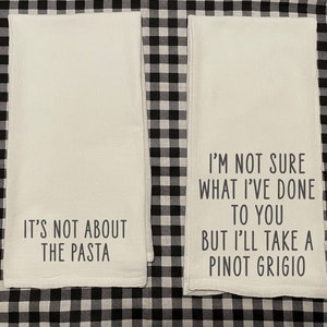 May include: Two white kitchen towels with black text. The first towel says "It's not about the pasta". The second towel says "I'm not sure what I've done to you but I'll take a Pinot Grigio".